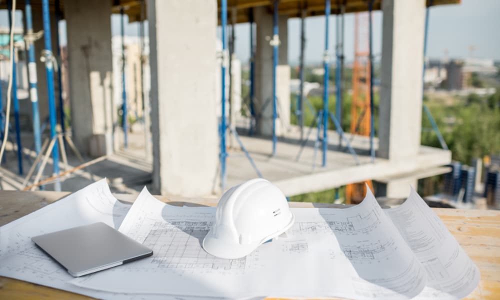 Construction plans on a table at a worksite with a hard hat and laptop laid on top.
