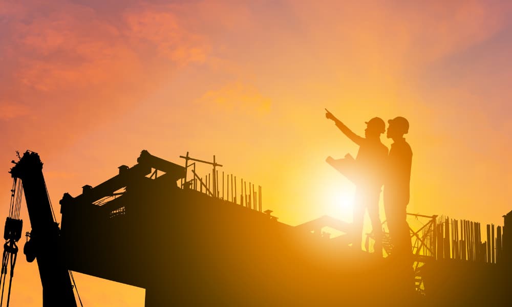Silhouette of two construction workers onsite against a rising sun, signalling skyward