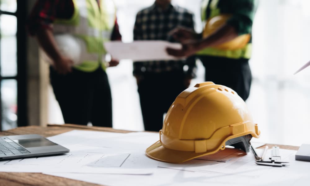 A yellow hard hat and a laptop are on a wooden table along with design paperwork and drafting instruments. Three members of a construction team are holding a discussion in the background.