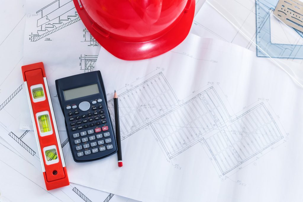 A top-down view of engineering tools and a red hard hat resting on architectural drawings.