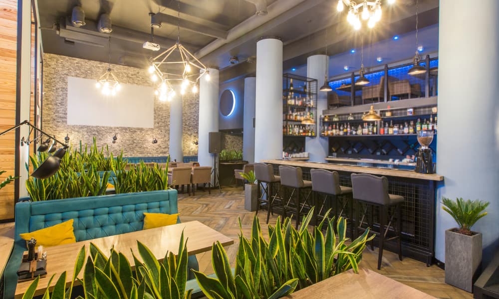 The interior of a modern restaurant. It is well-lit, has a bar area and table seating, and is decorated in warm natural tones of light wood, light stone, and green plants.