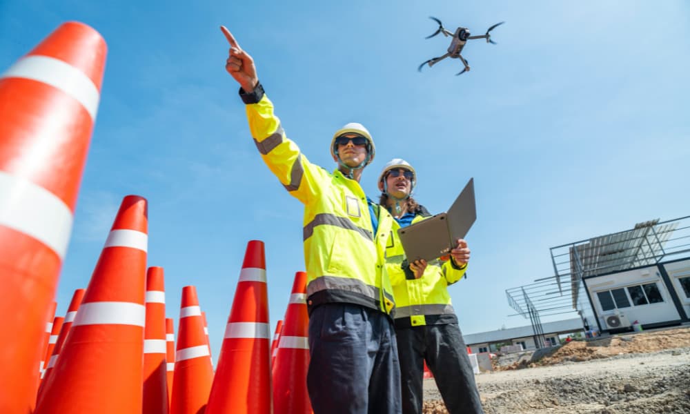 Two construction team members onsite wearing high-visibility jackets and hard hats. They are appraising the site using a laptop and a drone. There are a number of orange safety cones. It is a bright, cloudless day.